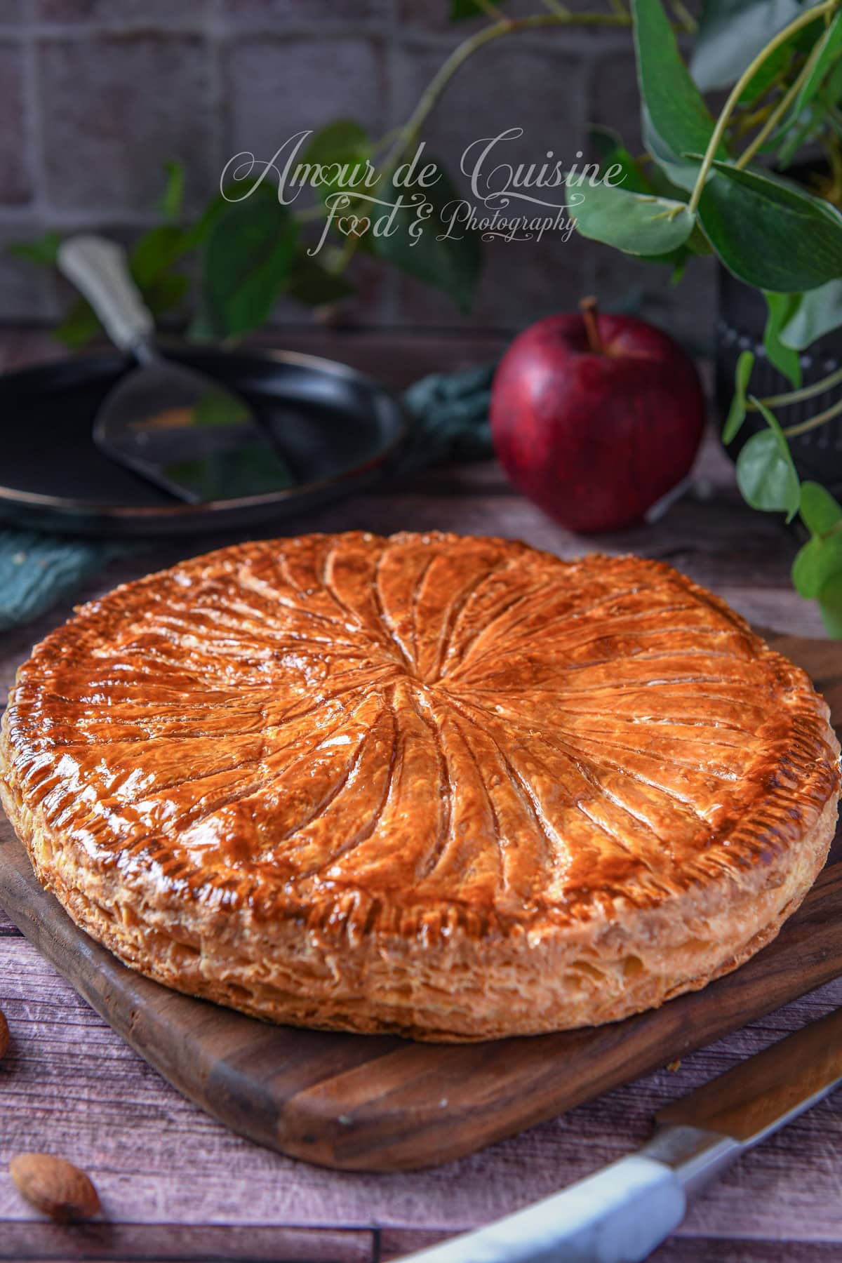 Whole Galette des Rois made with golden, glossy puff pastry, scored with a knife pattern, set on a wooden board; a spatula, a red apple, and blurred green leaves in the background.