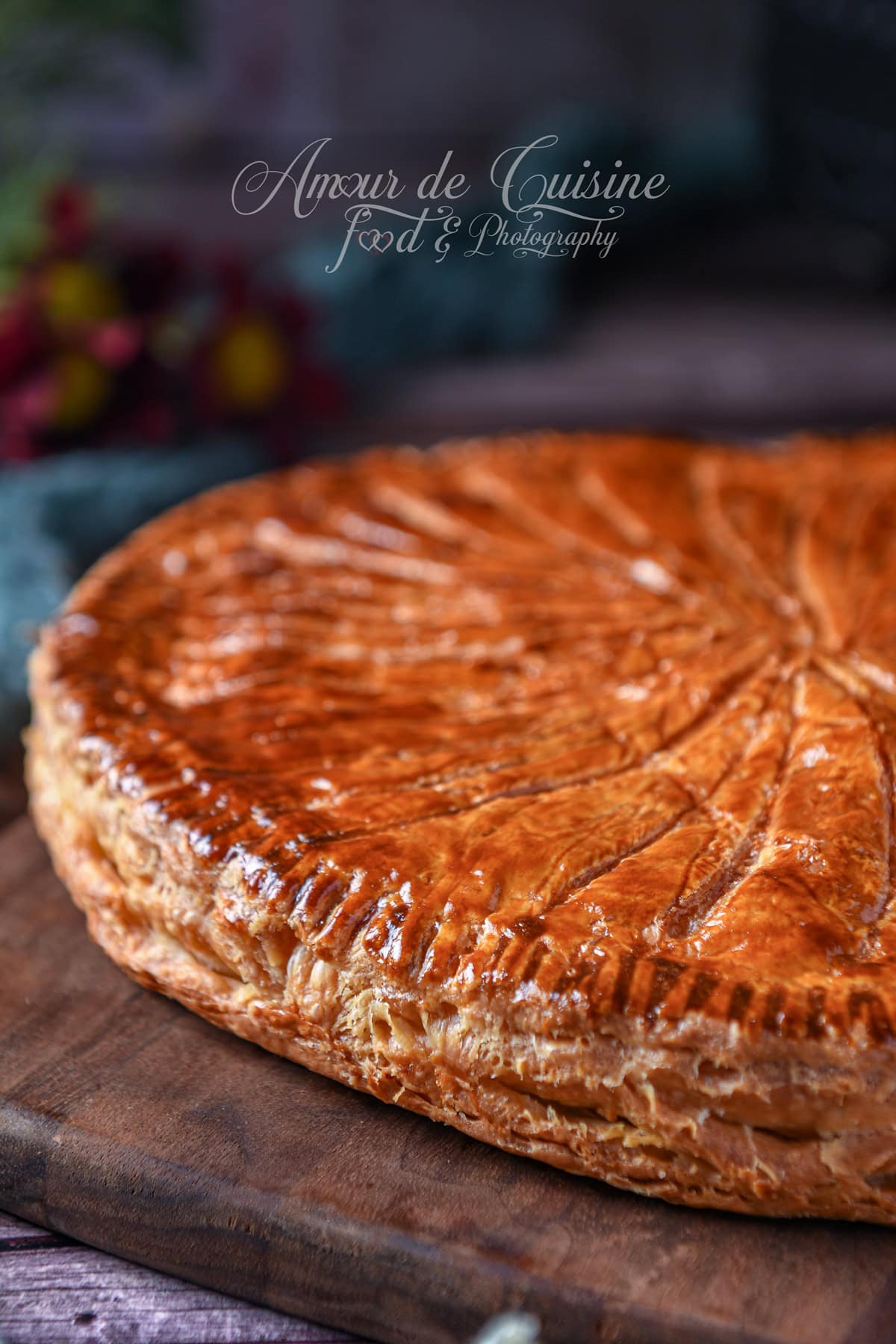 Close-up of a glossy, deeply golden puff pastry Galette des Rois, scored with a knife pattern, showing thick, crisp flaky layers on a wooden board.