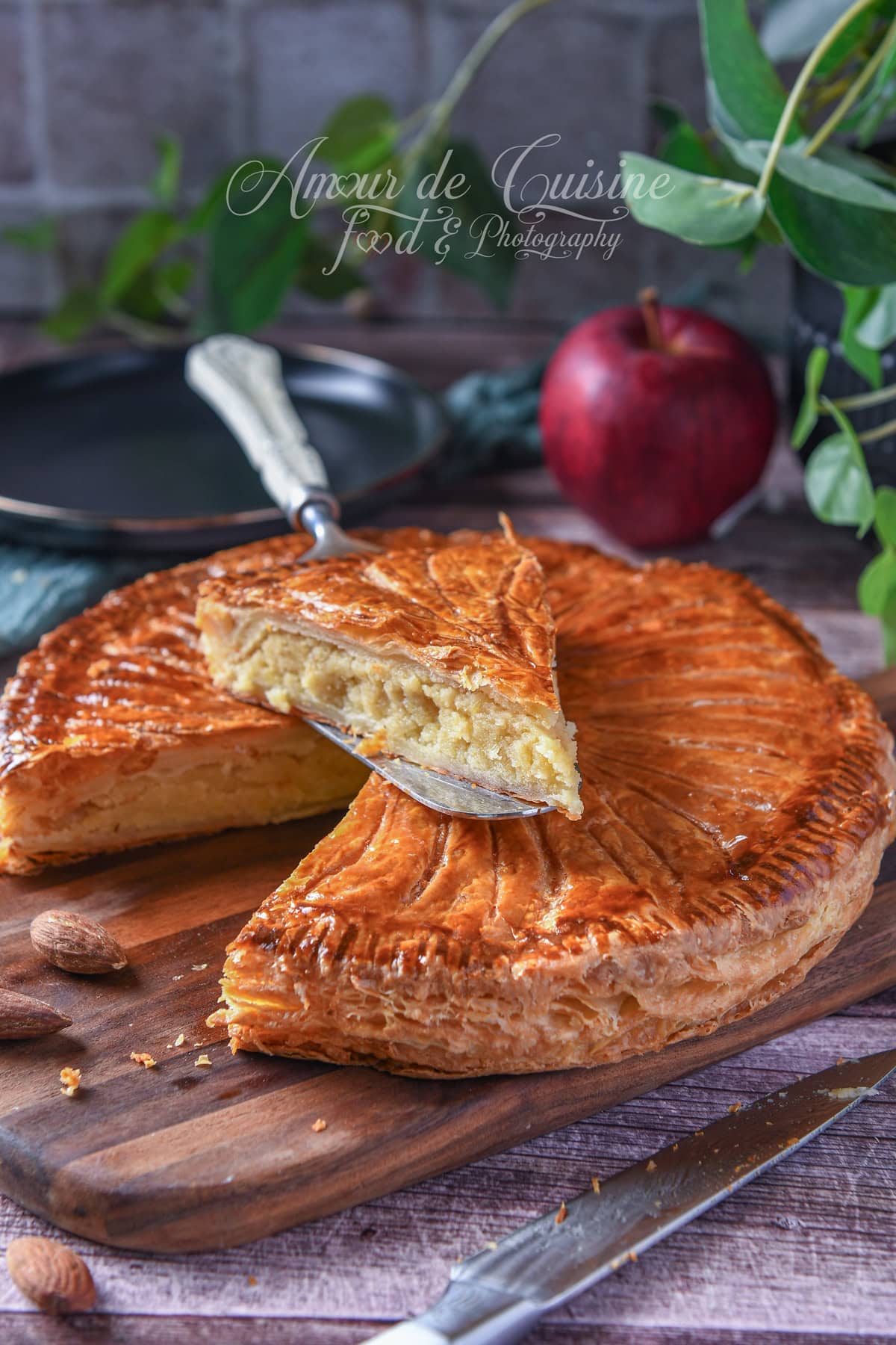 Golden puff pastry Galette des Rois on a wooden board, with a lifted slice showing the almond filling, a few almonds around, a knife in the foreground, and an apple in the background.