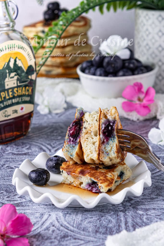 Fork slicing into a stack of fluffy blueberry pancakes on a white scalloped plate, drizzled with maple syrup and surrounded by fresh blueberries, with a bottle of syrup and more pancakes in the background.