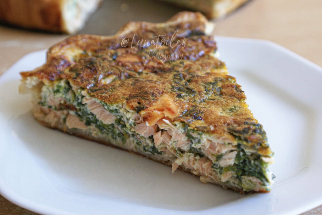 Part de quiche saumon épinard dorée, servie sur une assiette blanche, avec garniture fondante et morceaux visibles de saumon