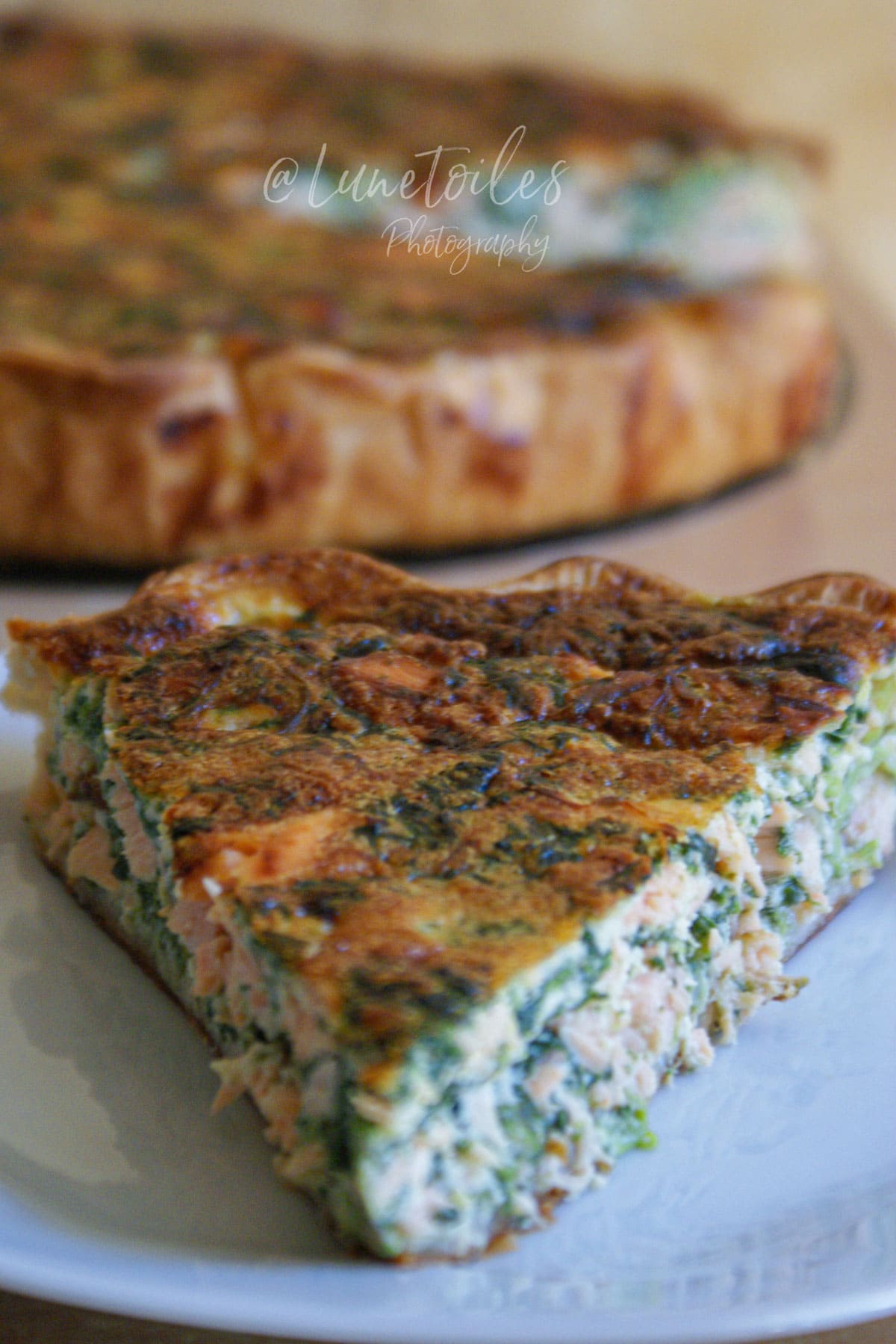 Part de quiche saumon épinard servie sur une assiette blanche, avec quiche entière floue en arrière-plan