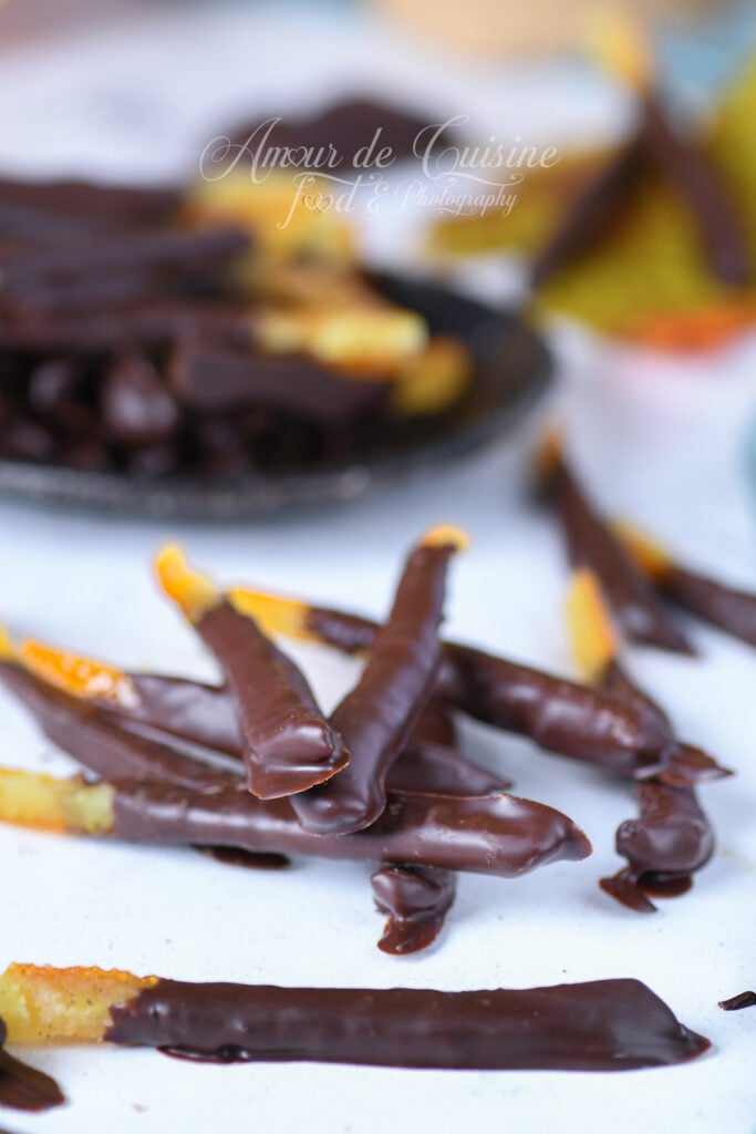 Homemade dark chocolate orangettes: candied orange peel sticks partly coated in chocolate, arranged on a light surface with a blurred bowl of orangettes in the background.