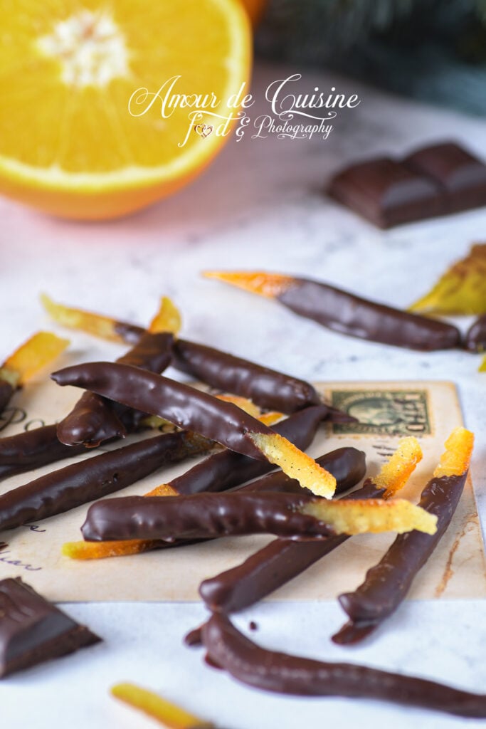 Homemade dark chocolate orangettes arranged on a vintage card, with a halved orange in the background and chocolate squares nearby.