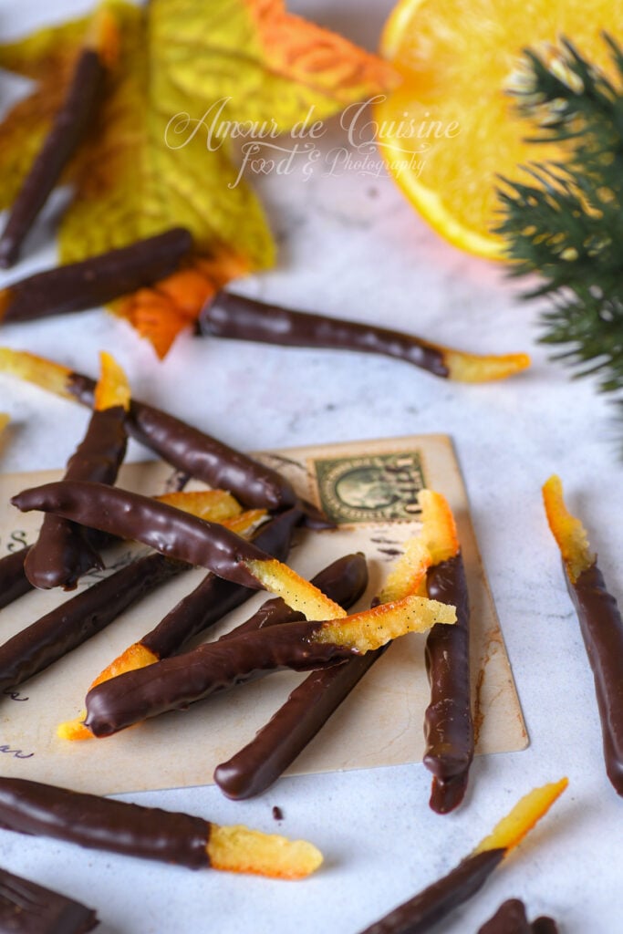 Homemade chocolate orangettes: candied orange peel sticks partly dipped in dark chocolate, arranged on a vintage card, with an orange slice and decorative leaves in the background.
