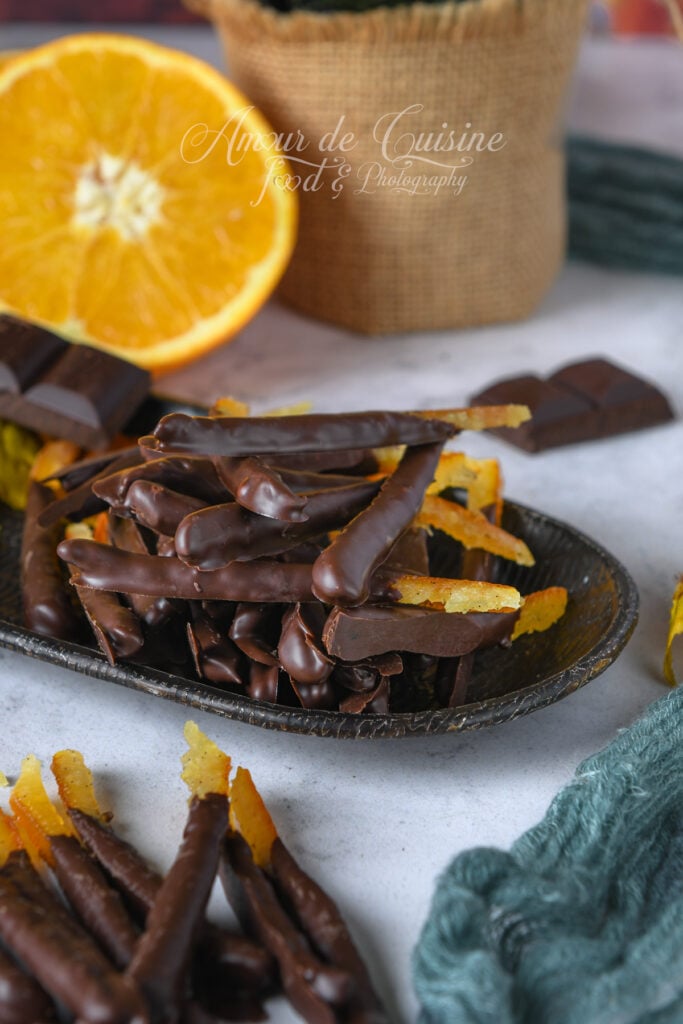 Black bowl of homemade dark chocolate orangettes, with a halved orange, chocolate squares, and a burlap bag in the background.