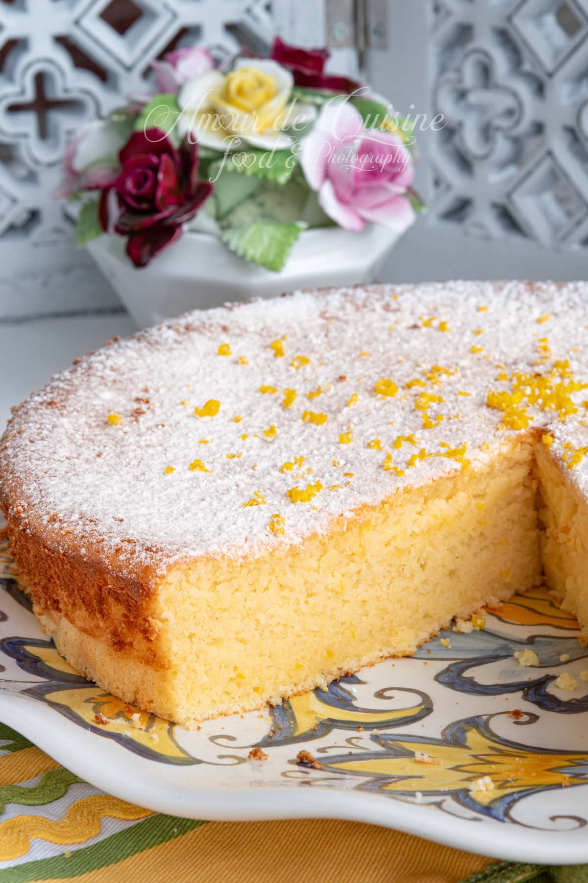 Close-up of a partially cut lemon sponge cake dusted with powdered sugar and sprinkled with lemon zest, served on a patterned platter with a blurred bouquet of flowers in the background.