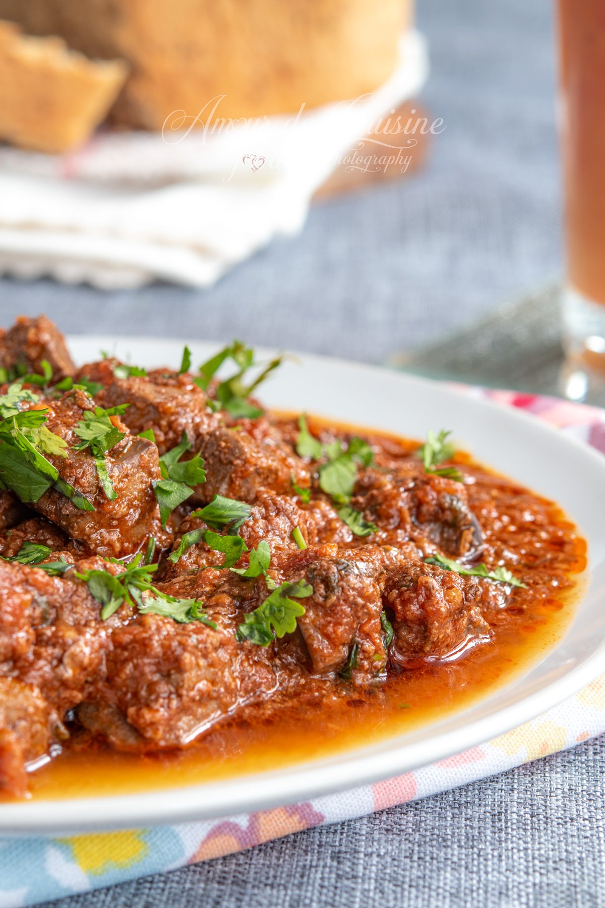 Close-up of kebda mchermla (lamb liver in spicy tomato sauce) with a glossy oil finish and fresh coriander on top, with homemade bread softly blurred in the background.