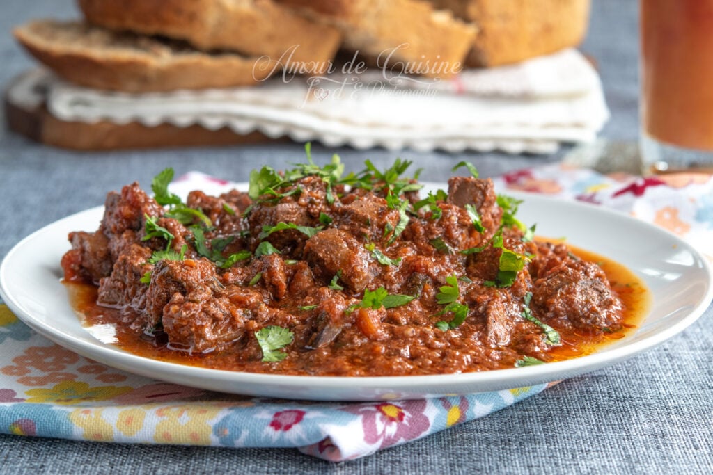 Plate of kebda mchermla (lamb liver in spicy tomato sauce) topped with fresh coriander, set on a floral cloth, with homemade bread and a fruity drink in the background.