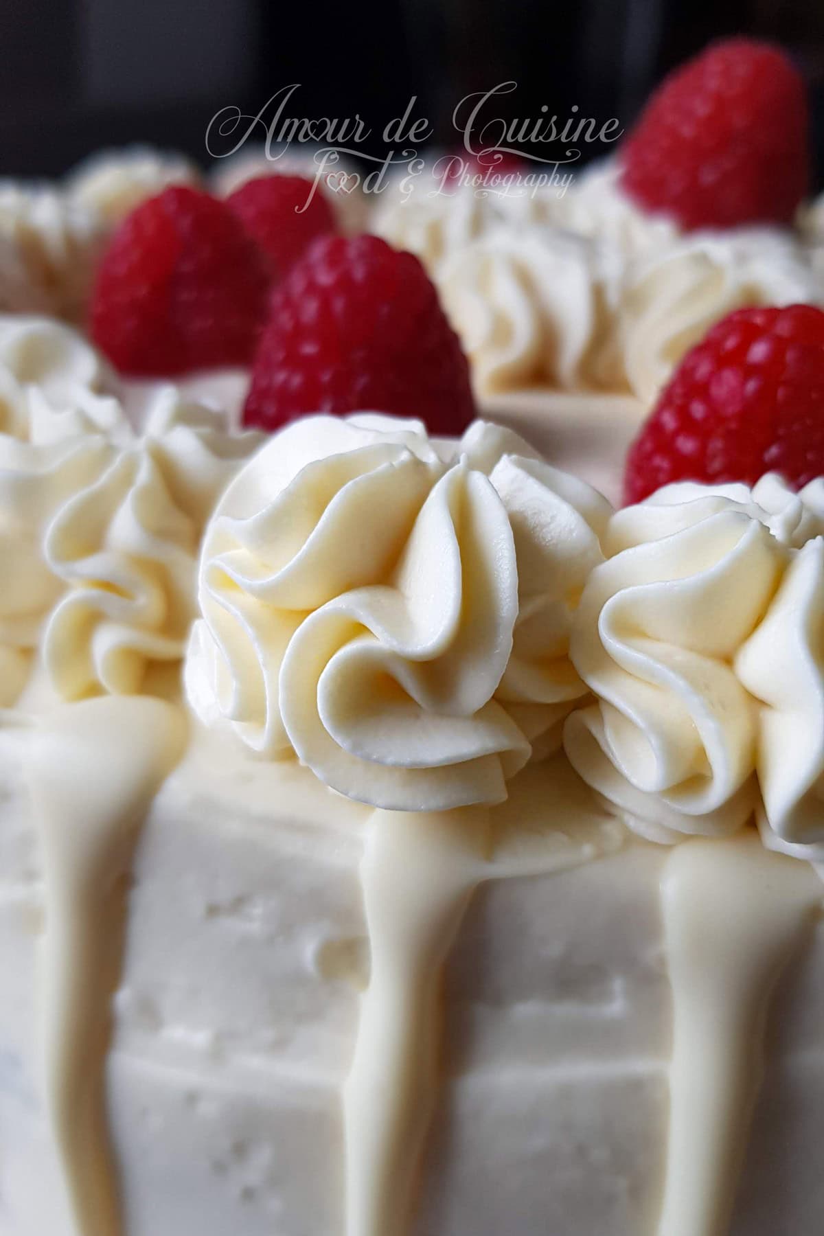 Gros plan d’un gâteau recouvert de ganache montée chocolat blanc et mascarpone, pochée en rosaces, avec coulis/glaçage blanc coulant et framboises fraîches sur le dessus.