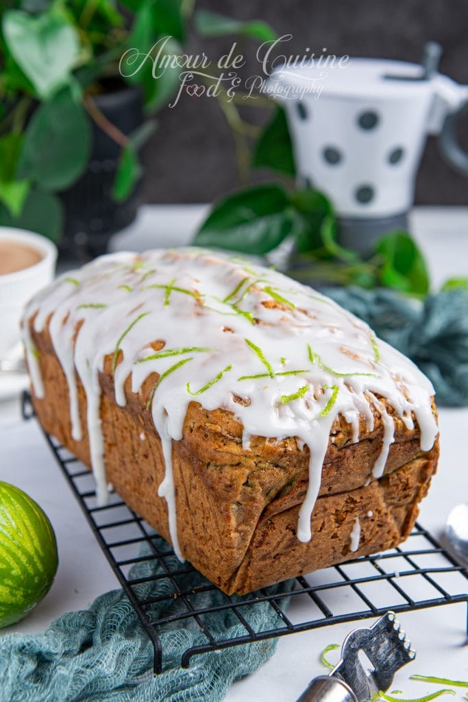 Gâteau aux courgettes entier, recouvert d’un glaçage blanc au citron vert et décoré de zestes, posé sur une grille de refroidissement avec un citron vert et des ustensiles en arrière-plan