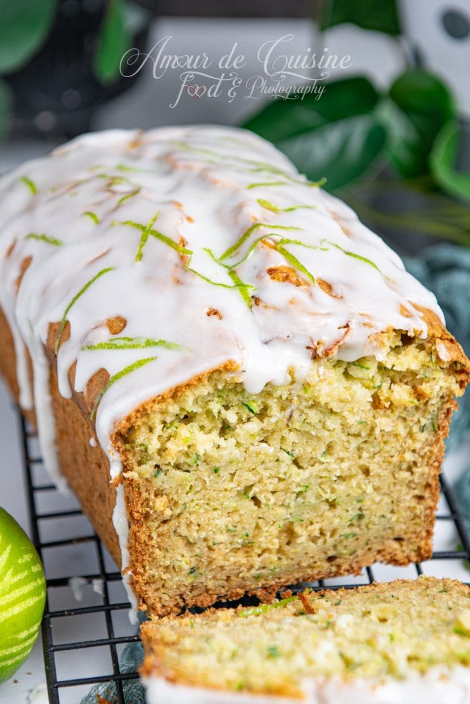 Gâteau moelleux aux courgettes nappé d’un glaçage au citron vert, présenté sur une grille de refroidissement, avec des zestes de citron vert en décoration et une tranche coupée au premier plan