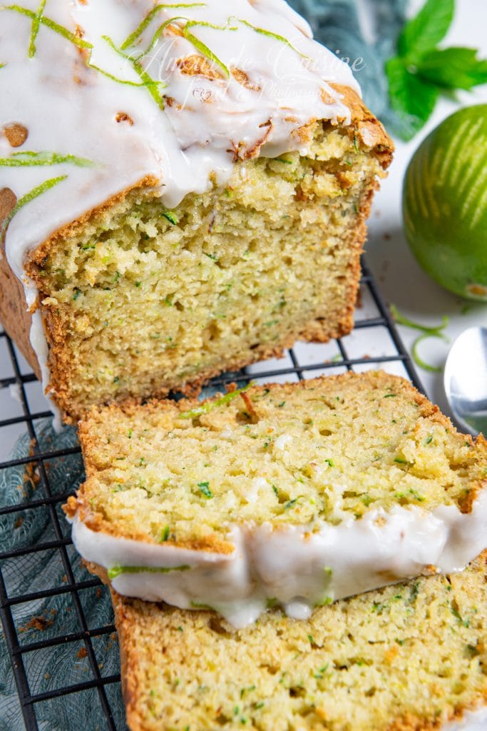 Tranche de gâteau aux courgettes et citron vert coupée et servie, montrant une mie moelleuse parsemée de courgettes râpées, surmontée d’un glaçage blanc brillant et décorée de zestes de citron