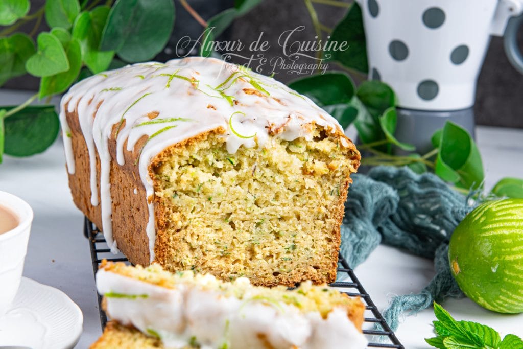 Gros plan sur un gâteau moelleux aux courgettes nappé d’un glaçage au citron vert, partiellement tranché, avec des zestes en décoration, posé sur une grille et entouré d’éléments verts et d’un citron