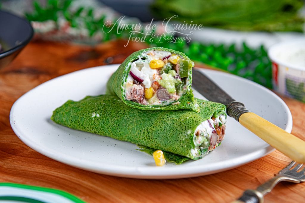 Wrap d’épinards coupé en deux, garni d’une farce fraîche à base de thon, maïs, avocat, oignon rouge, haricots et fromage crémeux, présenté sur une assiette blanche avec un couteau et une fourchette sur une table en bois