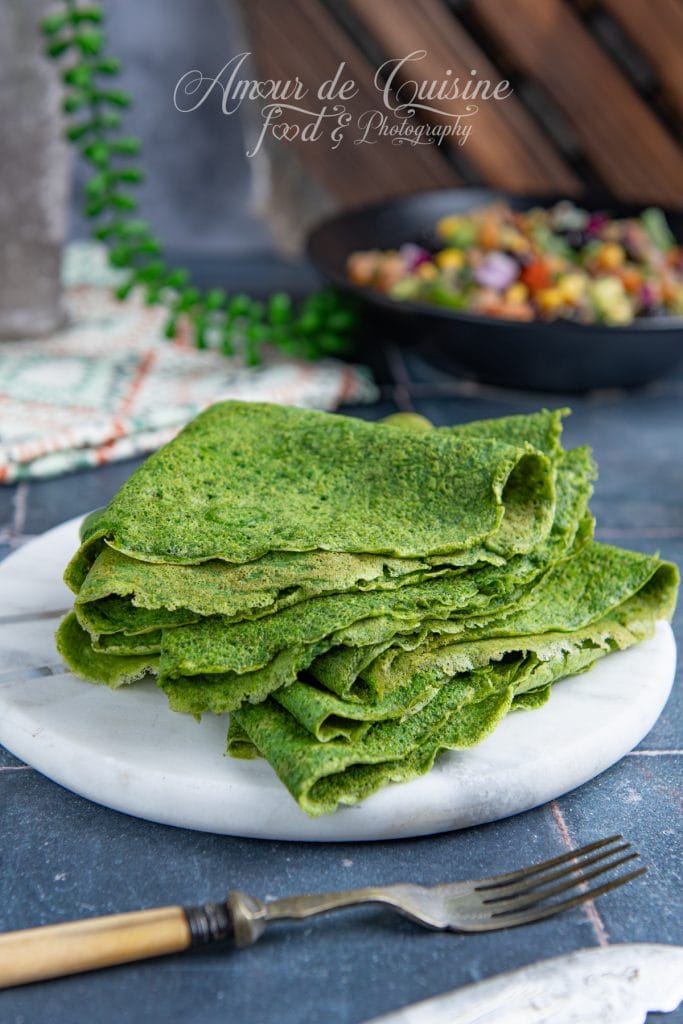 Pile de tortillas vertes aux épinards, soigneusement pliées et disposées sur une assiette en marbre blanc, avec une fourchette rustique au premier plan et une garniture colorée en arrière-plan flou