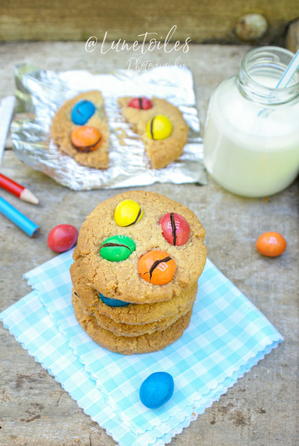 Cookies maison aux M&M’s colorés posés sur une serviette vichy bleue, accompagnés d’un verre de lait et de crayons de couleur, ambiance goûter enfantin et chaleureux – photo signée Lunetoiles