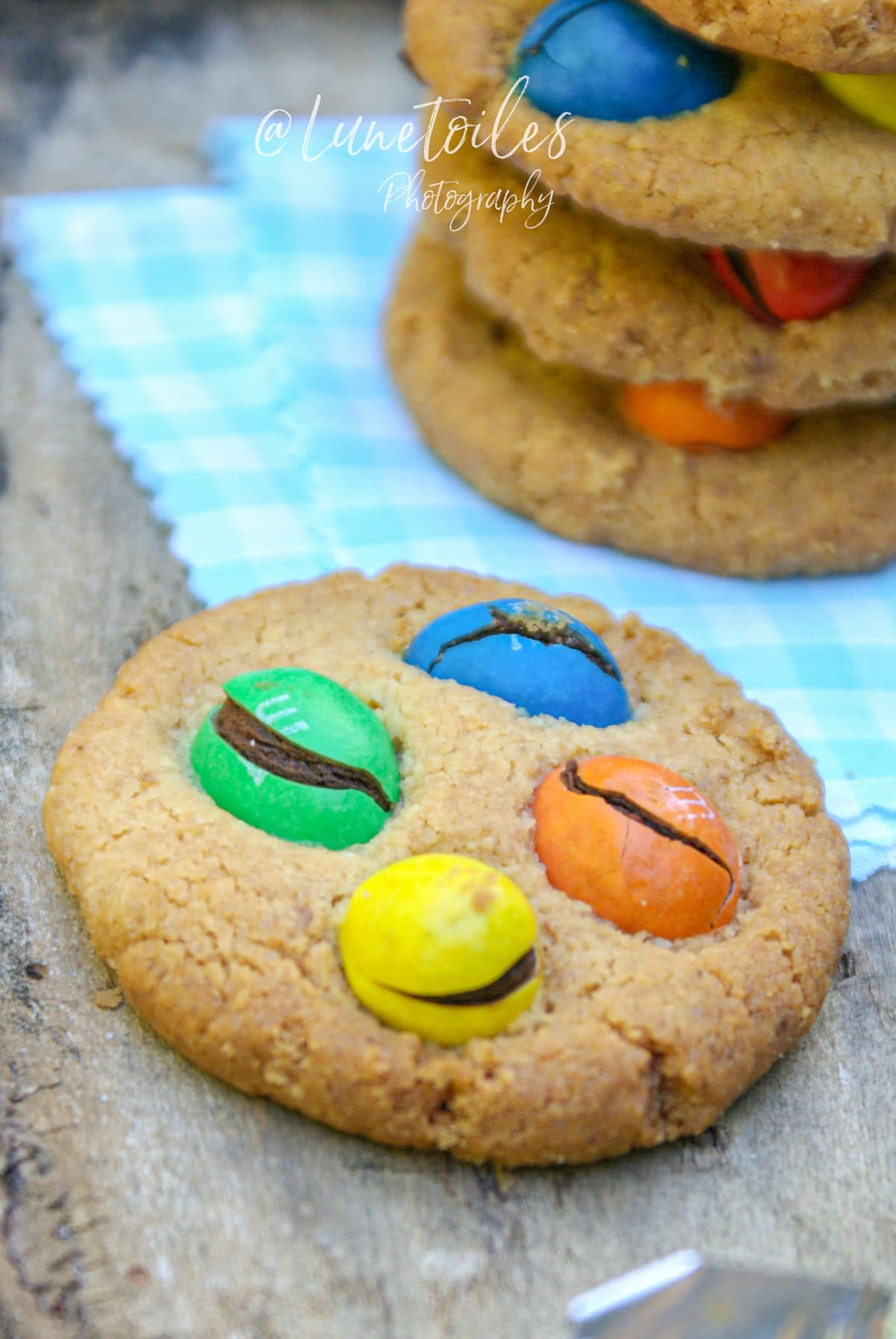 Cookie moelleux aux M&M’s colorés (jaune, vert, bleu, orange) bien craquelés, posé sur un fond en bois avec une serviette vichy bleue en arrière-plan – ambiance gourmande et fait maison