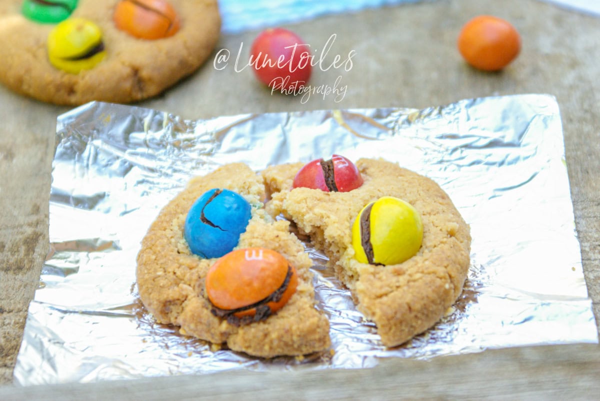 Cookie aux M&M’s cassé en deux, posé sur une feuille de papier aluminium, avec ses bonbons colorés (bleu, rouge, jaune, orange) légèrement craquelés – ambiance goûter fait maison, photo signée Lunetoiles.