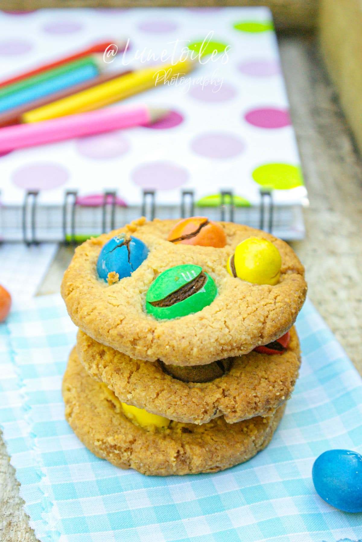 Cookies dorés aux M&M’s colorés empilés sur une serviette vichy bleue, posés devant un cahier à pois et des crayons – une pause gourmande signée Lunetoiles