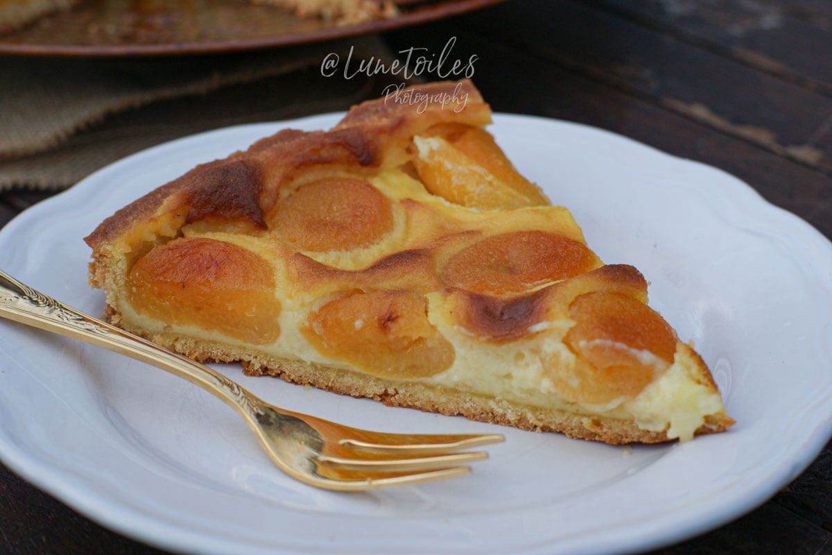 Part de tarte aux abricots à la crème pâtissière servie sur une assiette blanche, avec une pâte sucrée dorée