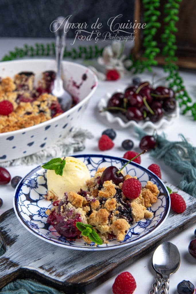 Assiette de crumble d'amandes aux fruits rouges servi avec une boule de glace vanille, décorée de feuilles de menthe, framboises et cerises fraîches, posée sur un plateau en bois avec en arrière-plan un plat de cuisson et des fruits rouges éparpillés