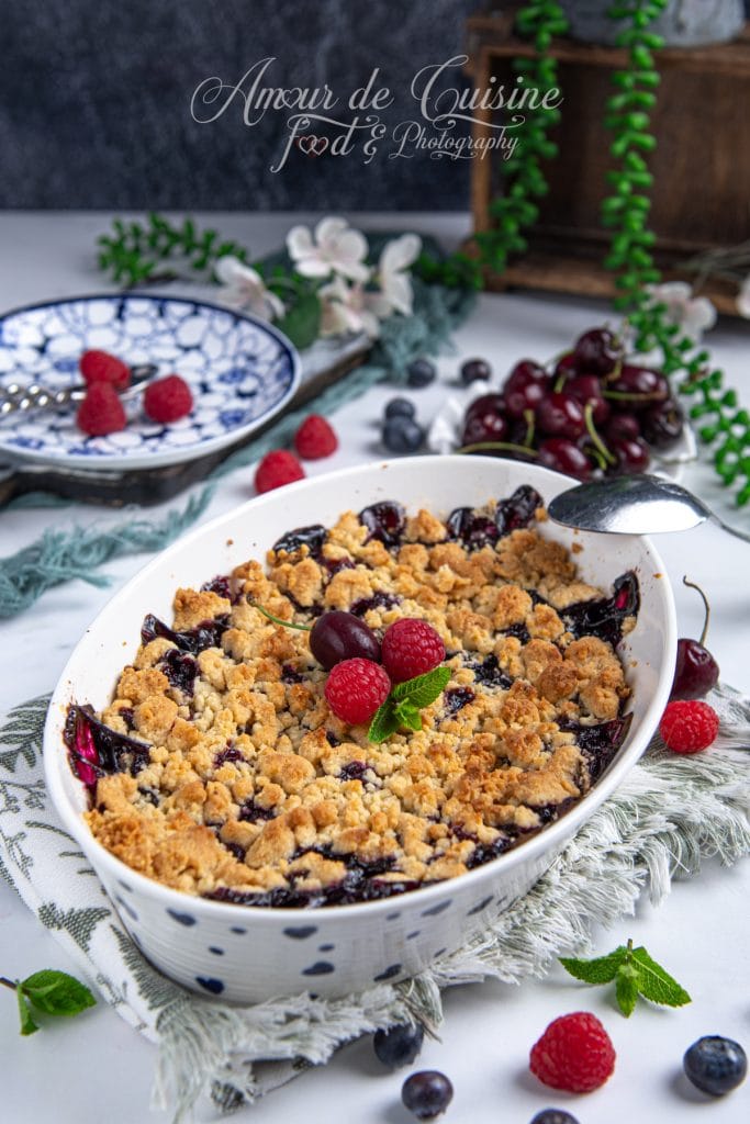 Plat de crumble d'amandes aux fruits rouges fraîchement sorti du four, garni de framboises, cerise et menthe fraîche, entouré de fruits rouges et décorations florales sur une table blanche et élégante