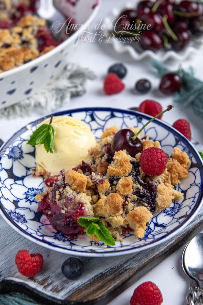 Portion de crumble aux fruits rouges servie dans une assiette fleurie avec une boule de glace vanille, décorée de menthe fraîche, framboise et cerise, sur un plateau en bois entouré de fruits rouges.