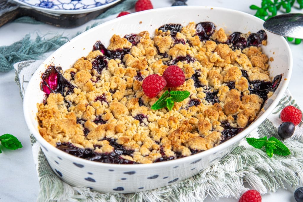 Plat de crumble aux fruits rouges fraîchement cuit, doré et croustillant, garni de framboises fraîches et de feuilles de menthe, présenté sur un torchon en lin avec quelques fruits rouges autour