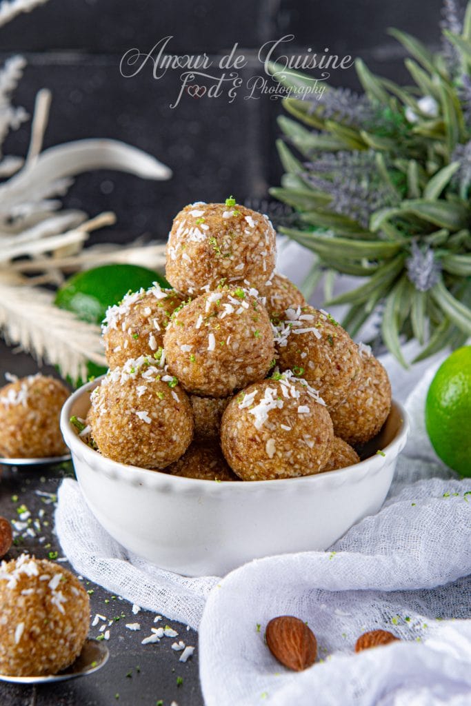No-bake almond lime energy balls topped with shredded coconut in a white bowl, surrounded by limes and almonds, on a dark surface.