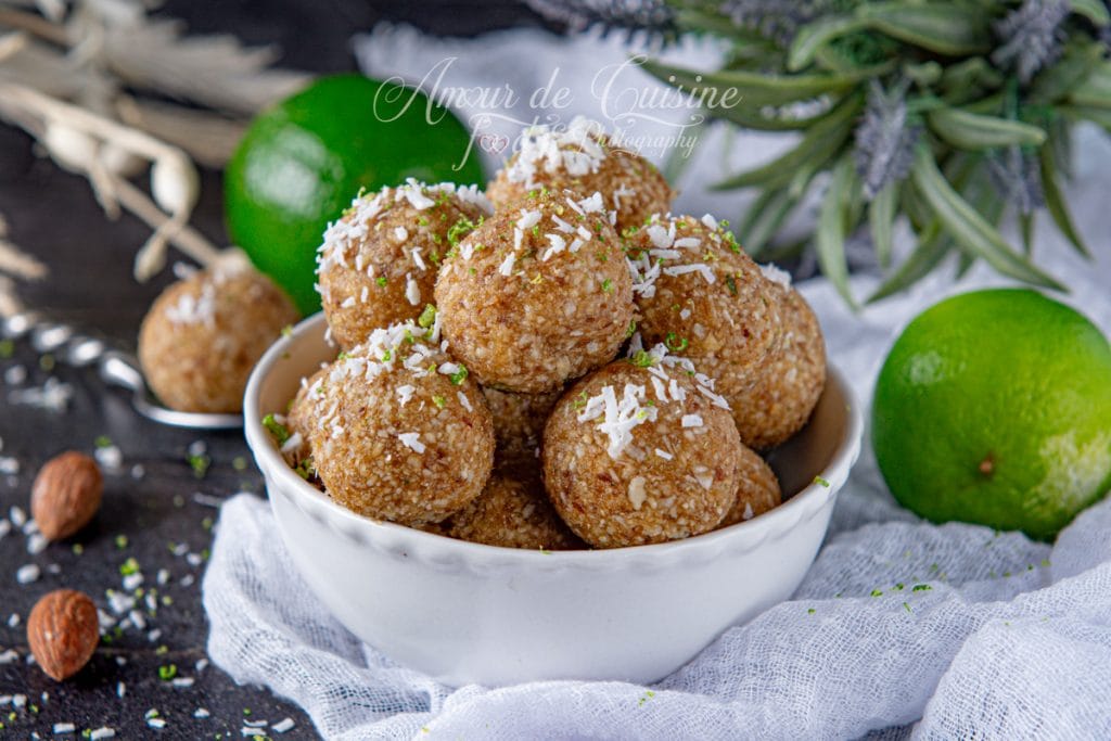 Homemade energy bites with almonds, dates, and lime, served in a white bowl, topped with shredded coconut, and styled with whole limes and almonds on a dark background