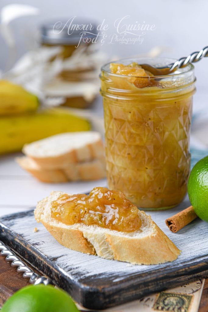 Confiture de banane au citron vert maison servie sur une tranche de pain frais, avec bocal en verre rempli de confiture, citron vert et bâton de cannelle en arrière-plan