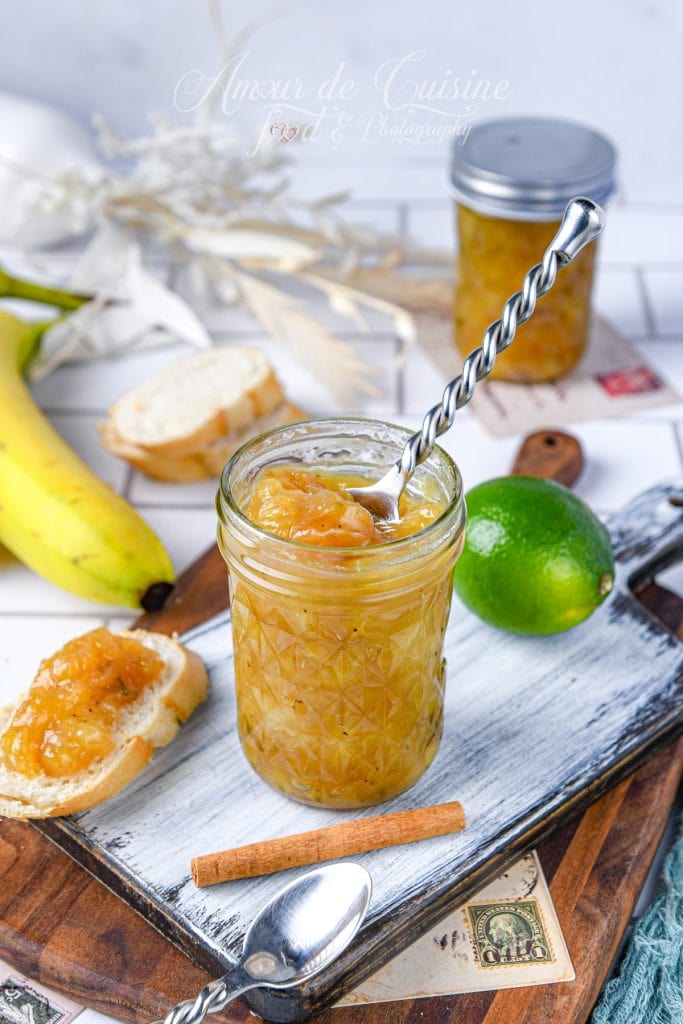 Confiture de banane au citron vert maison dans un bocal en verre, avec cuillère et tranche de pain tartinée, accompagnée d’une banane, d’un citron vert et d’un bâton de cannelle.