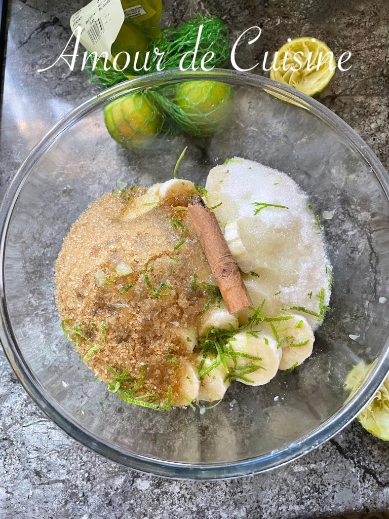 Ingrédients de la confiture de banane au citron vert dans un saladier : rondelles de banane, sucre blanc et roux, zeste de citron vert râpé et bâton de cannelle prêts à être mélangés.