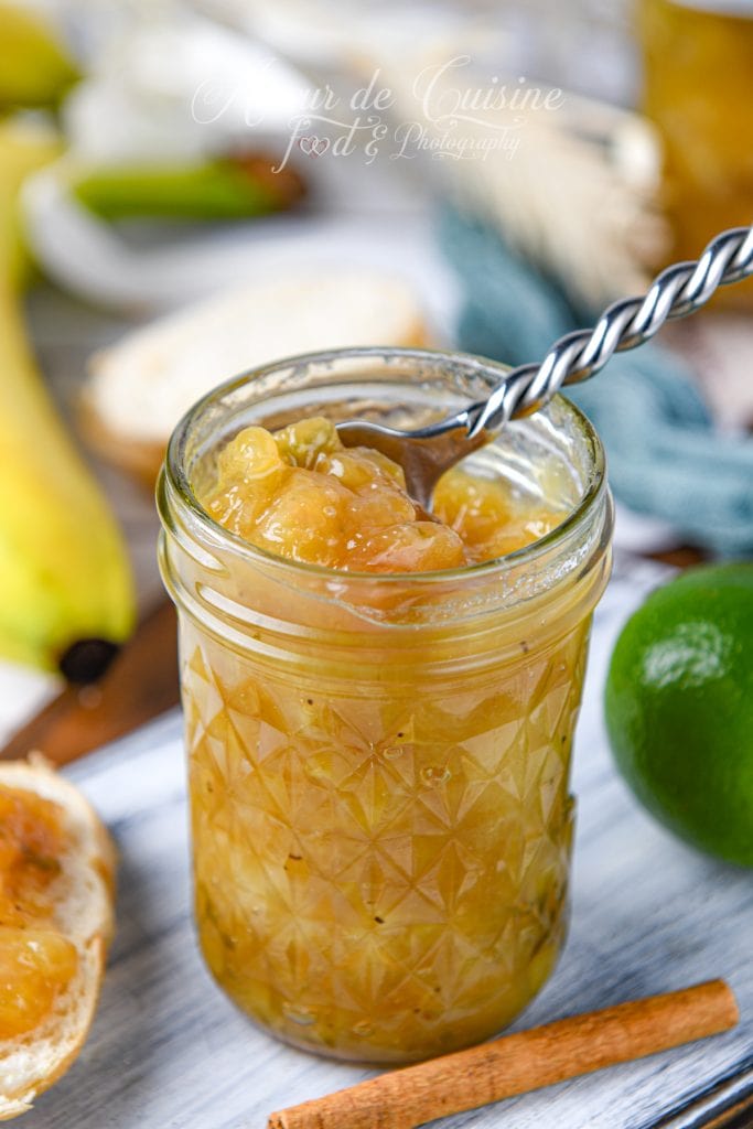 Bocal en verre rempli de confiture de banane au citron vert maison, avec une cuillère et des morceaux de banane visibles, accompagné d’un citron vert, d’un bâton de cannelle et d’une tranche de pain tartinée.