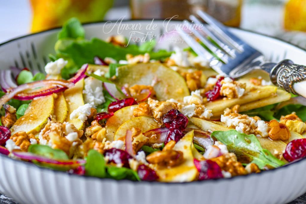 Gros plan sur une salade aux poires, roquette, feta émiettée, noix de pécan croquantes et cranberries séchées, arrosée de vinaigrette balsamique-érable et servie dans un bol blanc avec une fourchette élégante.