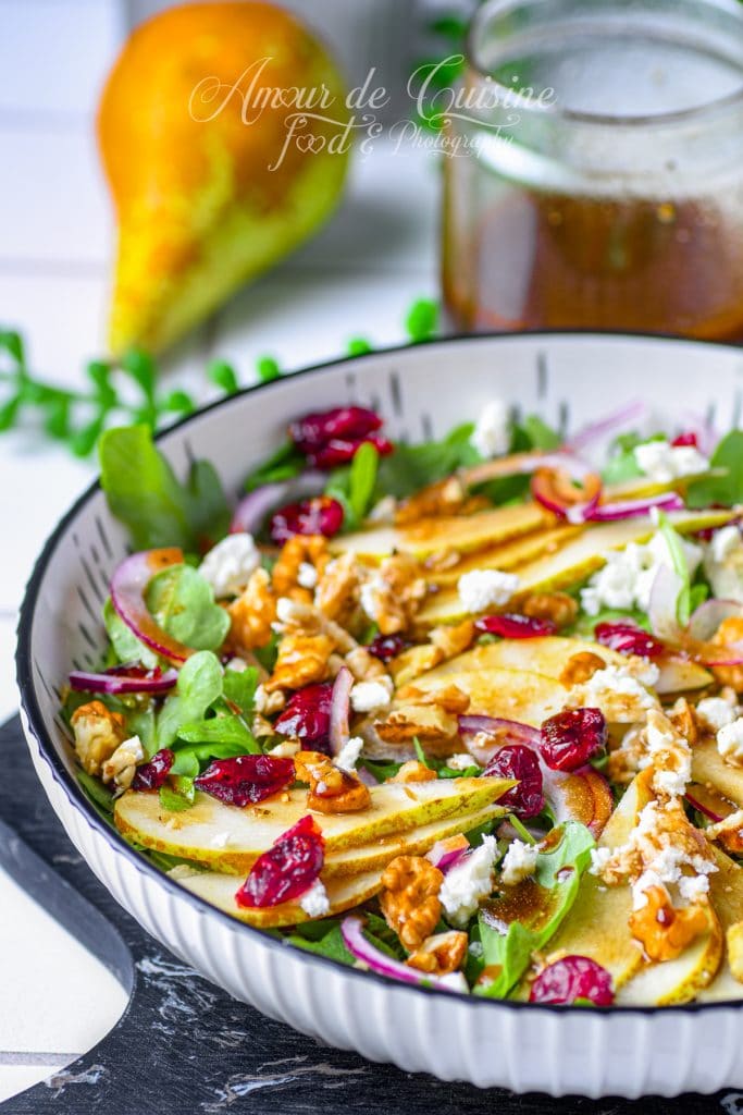 Salade aux poires, roquette, feta, noix de pécan et cranberries séchées dans un bol, garnie d’oignon rouge et nappée de vinaigrette balsamique-érable maison, avec une poire fraîche en arrière-plan.