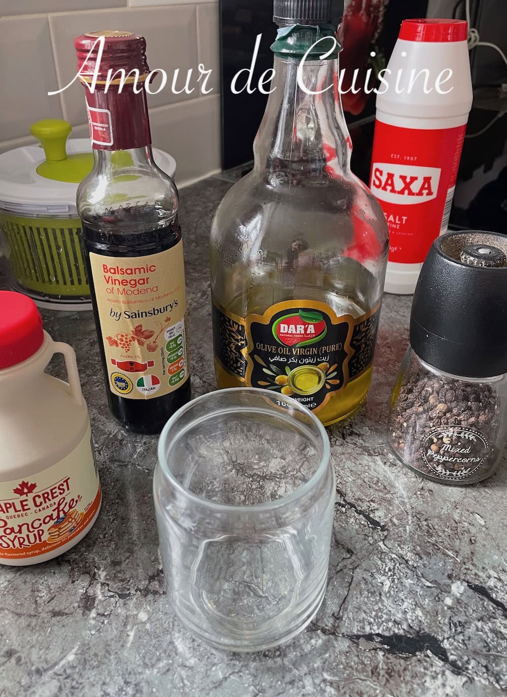 Ingrédients pour vinaigrette balsamique à l’érable sur un plan de travail : bouteille de vinaigre balsamique, huile d’olive, sirop d’érable, sel, moulin à poivre et bocal en verre.