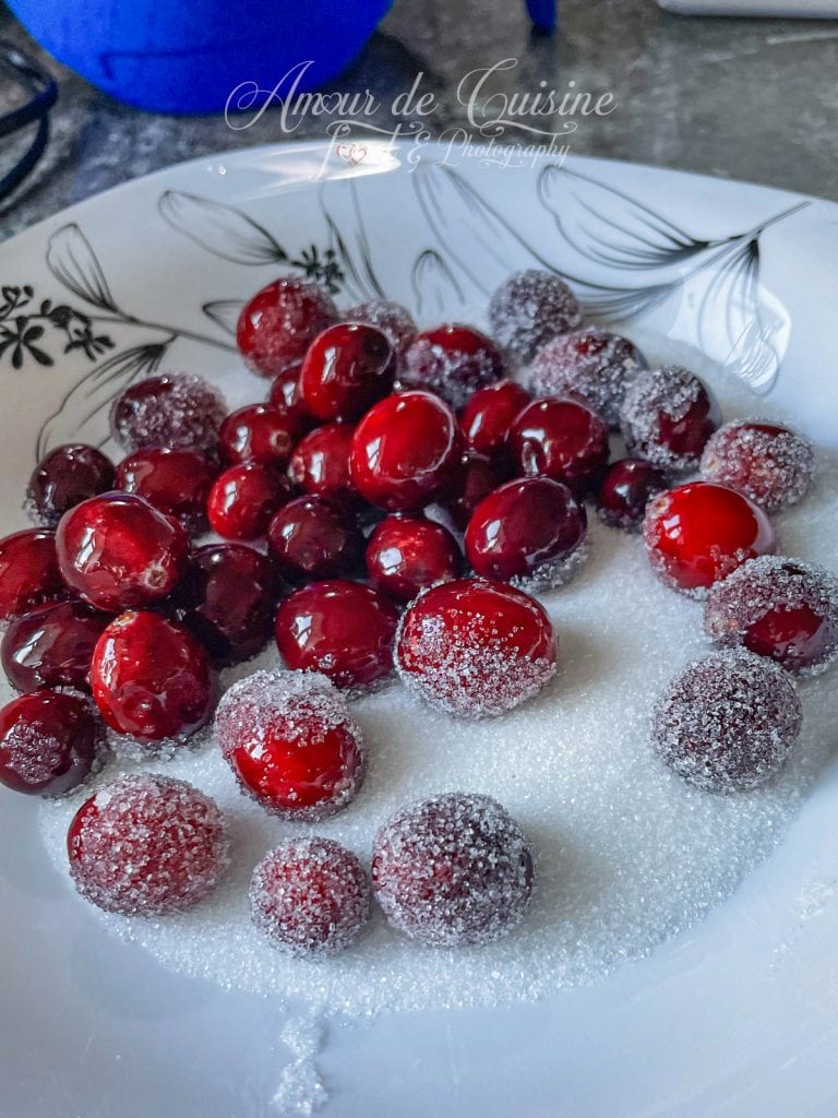 Cranberries sucrées givrées disposées sur une assiette en bois et parsemées de sucre cristallisé, avec en arrière-plan un bol contenant d'autres baies enrobées.