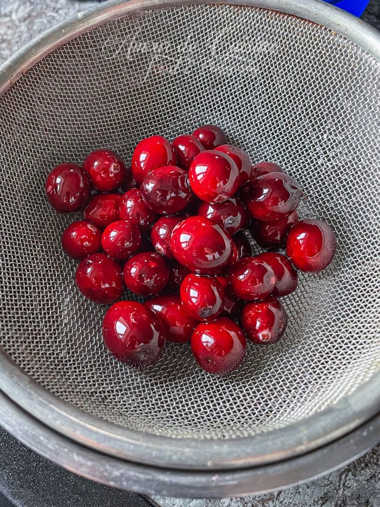 Canneberges fraîches et brillantes en train de s'égoutter dans une passoire fine après avoir été trempées dans un sirop, étape de préparation des cranberries sucrées givrées.