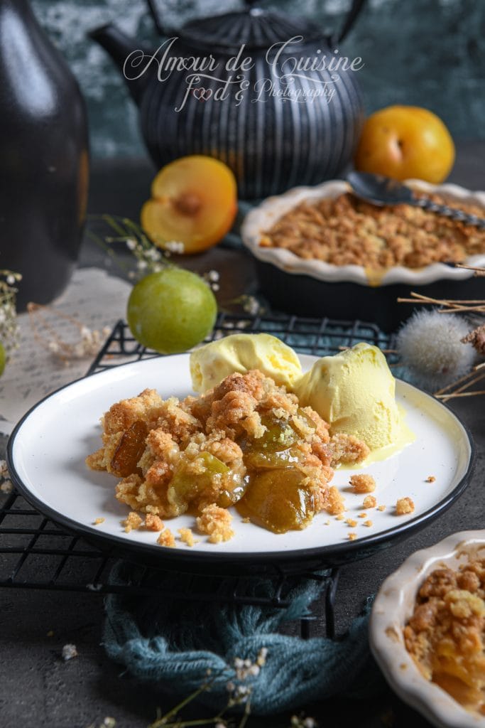 Assiette de crumble aux reines-claudes et mirabelles, servi tiède avec deux boules de glace vanille, en ambiance rustique avec prunes fraîches et plat de crumble en arrière-plan.