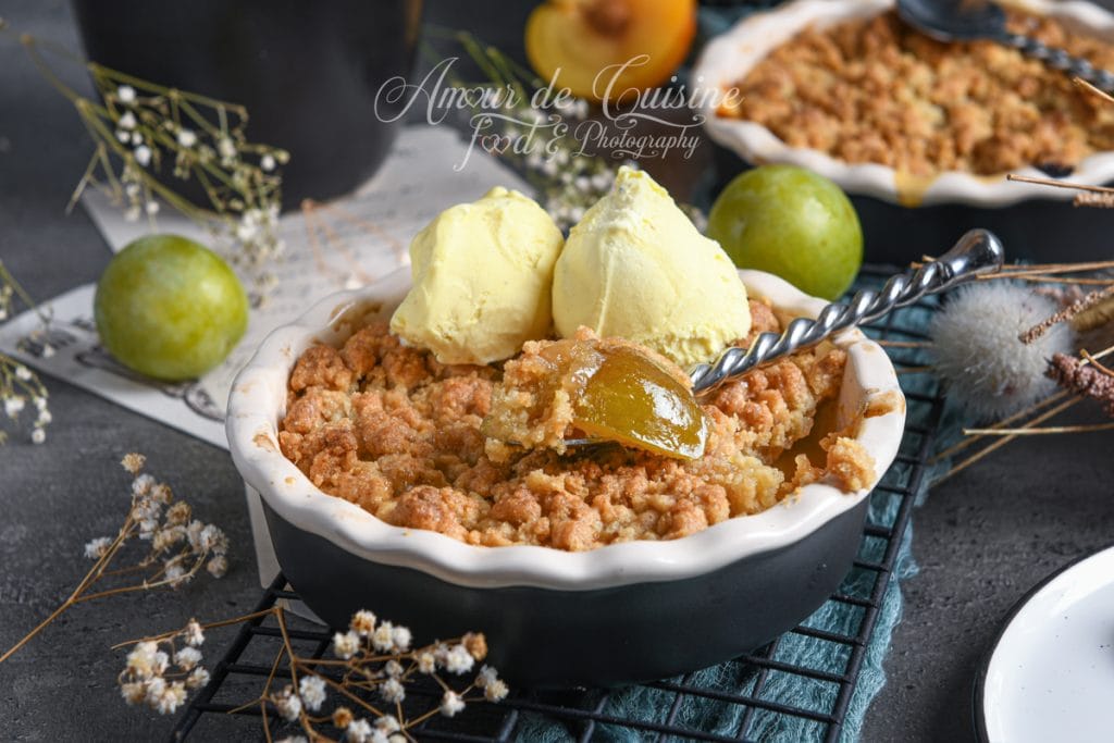 Crumble aux reines-claudes et mirabelles servi dans un ramequin, surmonté de deux boules de glace vanille, avec une cuillère plongeant dans des fruits fondants, entouré de prunes fraîches et décor floral.