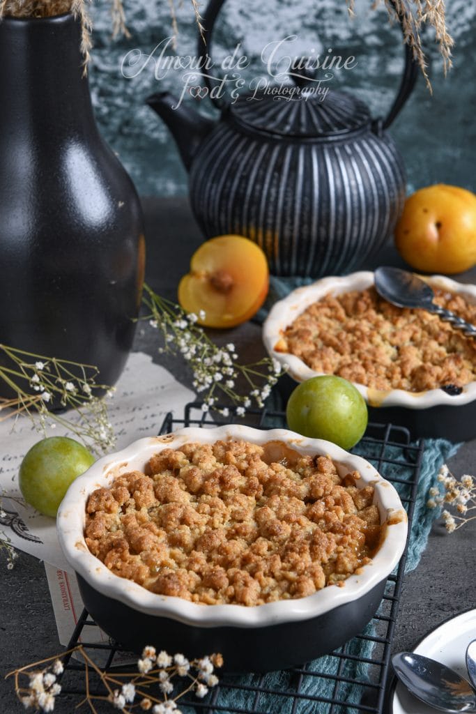 Crumble doré aux reines-claudes et mirabelles, présenté dans un plat à tarte individuel, entouré de prunes fraîches, d’accessoires rustiques et d’une théière noire en arrière-plan.