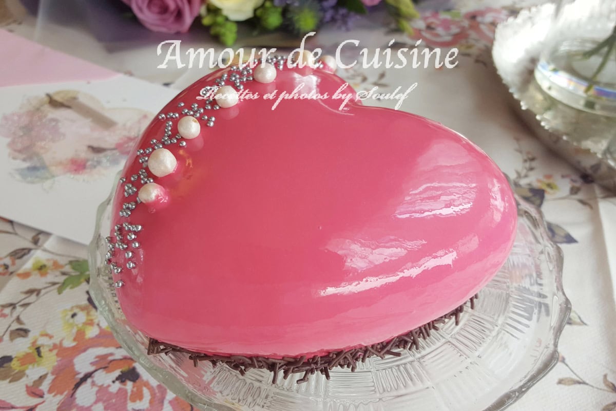 Heart-shaped strawberry entremet coated with a glossy pink mirror glaze, decorated with pearls and silver sprinkles, displayed on a glass stand with flowers in the background.
