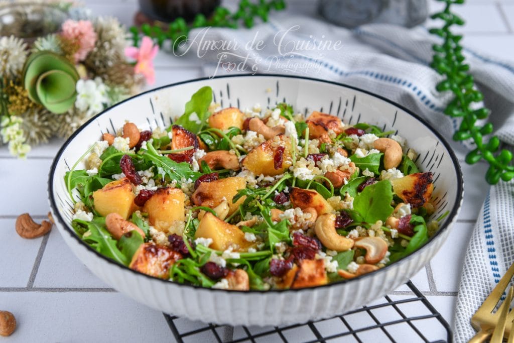 Salade colorée de coings rôtis, quinoa et roquette, garnie de feta émiettée, cranberries séchées et noix de cajou grillées, servie dans un grand bol sur une table décorée.