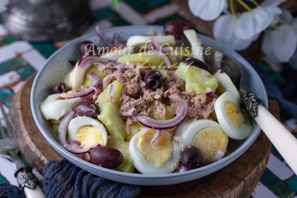 Bowl of potato and egg salad topped with tuna, red onion slices, and black olives, served on a rustic wooden board with elegant vintage cutlery and soft lighting for a homely feel.