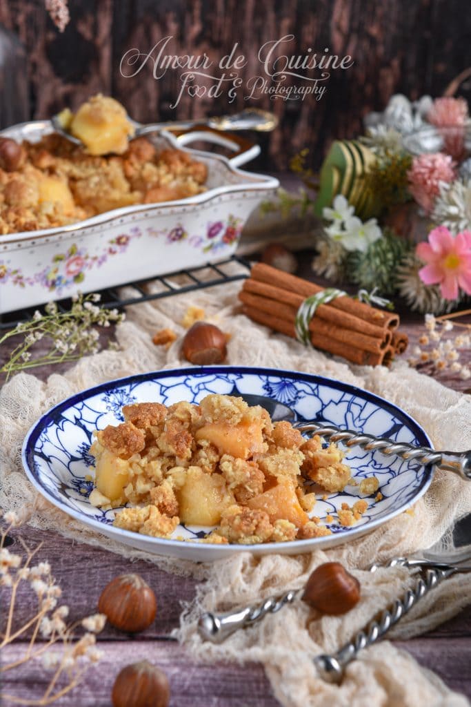 Crumble de coings aux noisettes doré et croustillant servi dans une assiette à motifs bleus, avec un plat de cuisson fleuri à l’arrière-plan, des noisettes entières et des bâtons de cannelle en décor rustique