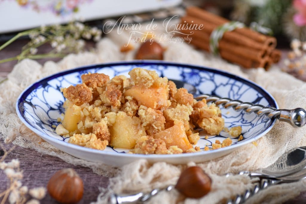 Portion de crumble de coings aux noisettes servie dans une assiette à motifs bleus, avec morceaux de fruits fondants et pâte dorée croustillante, décorée de noisettes et bâtons de cannelle en arrière-plan