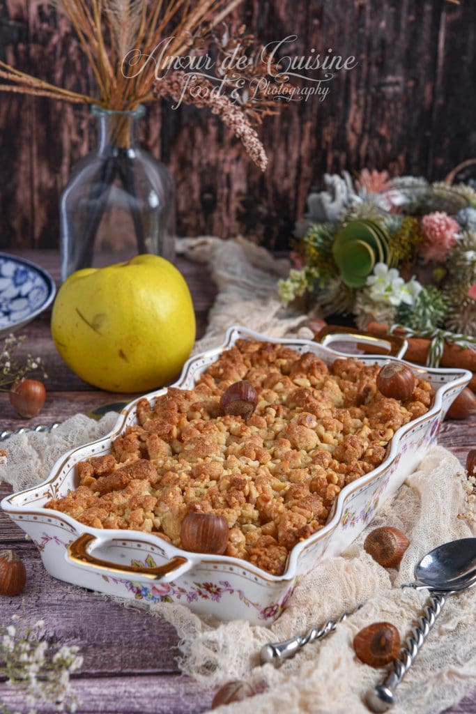 Crumble de coings aux noisettes doré à la sortie du four, présenté dans un plat à gratin fleuri, entouré de noisettes entières, d’un coing frais et d’une décoration automnale rustique.