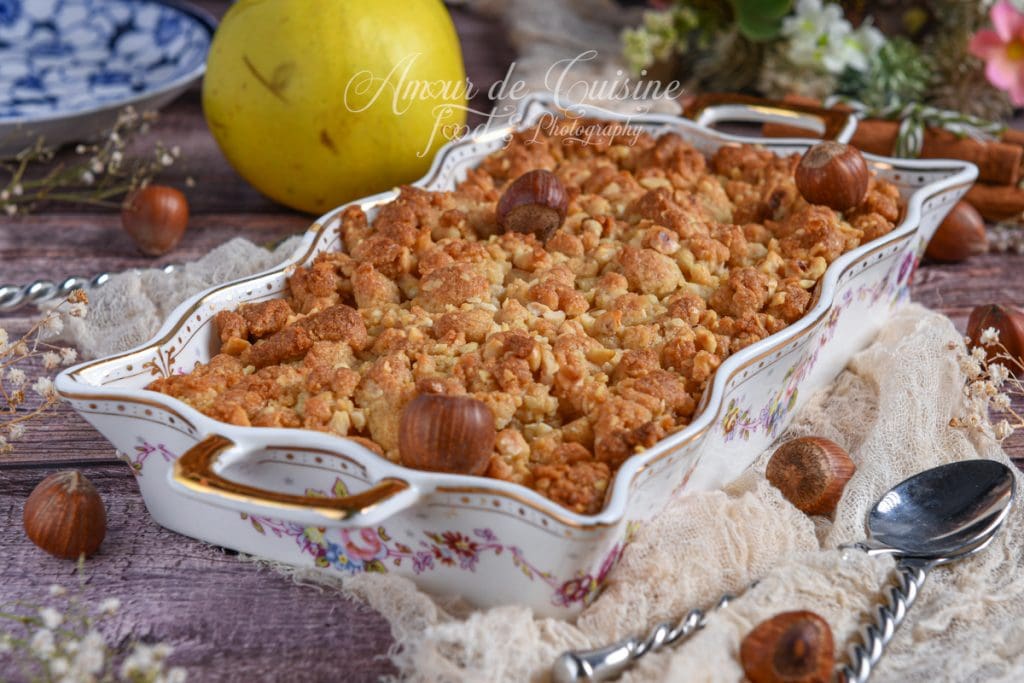 Crumble de coings aux noisettes doré et croustillant sorti du four, présenté dans un plat à gratin fleuri avec des noisettes entières en décor, sur une table rustique en bois